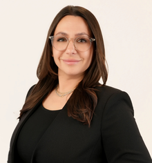 Professional portrait of a woman with long brown hair, glasses, and black blazer.