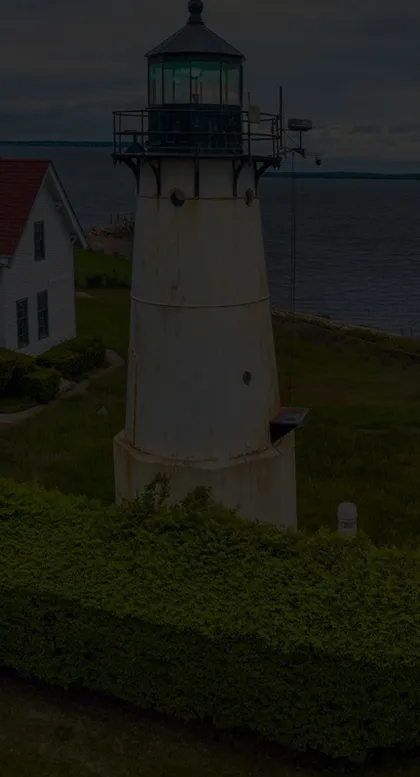 White cylindrical lighthouse with black railing and lantern room, adjacent to a white house with a red roof near a body of water.