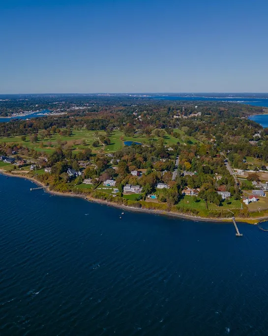 Aerial view of a coastal residential area with green lawns, trees, and blue water under a clear sky.