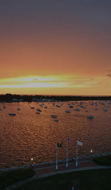 Sunset over a harbor with numerous boats anchored and three flags on poles in the foreground.