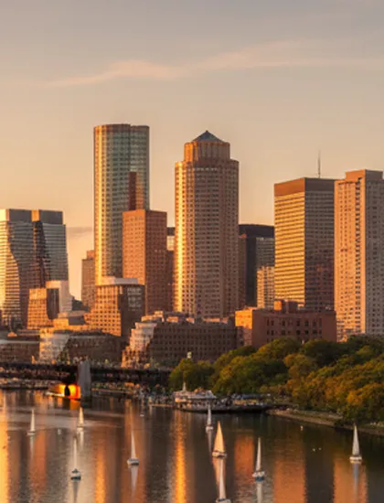 Downtown city skyline at sunset with tall buildings, river, and sailboats.