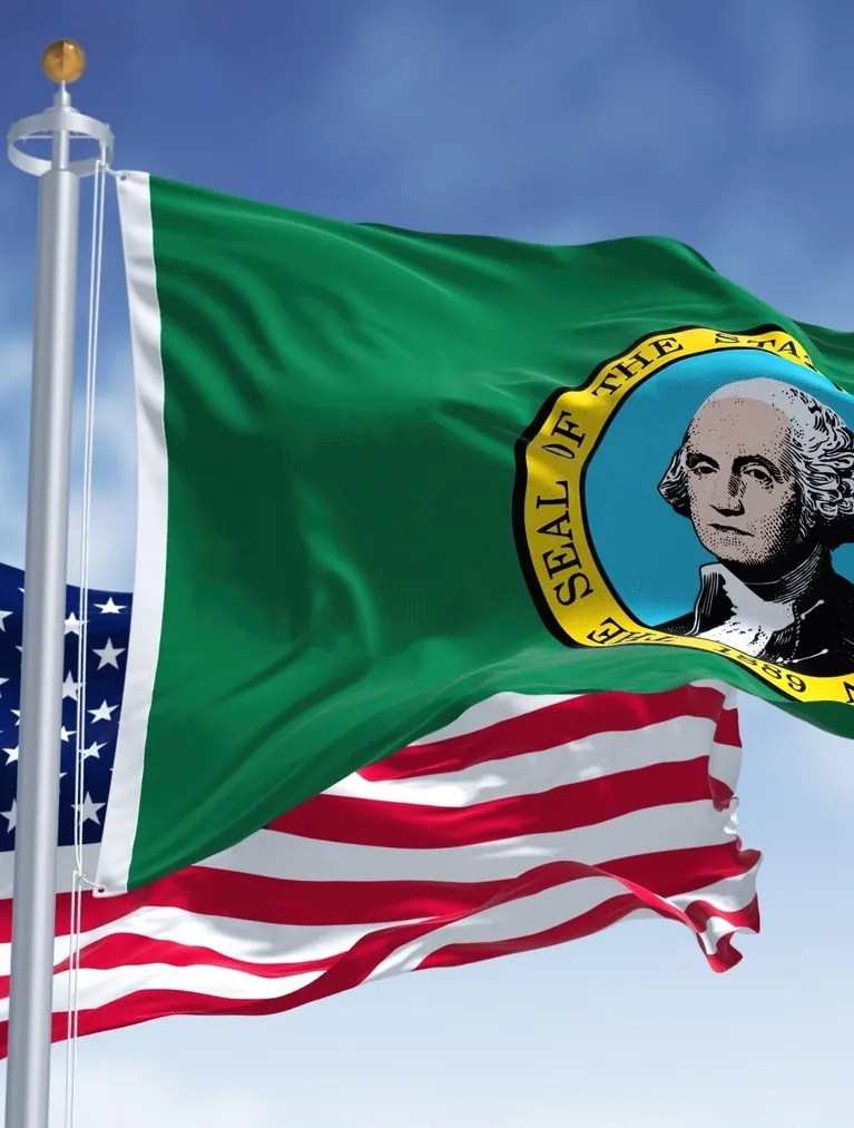 Two flags waving on flagpoles against a blue sky: the Washington state flag above the United States flag.
