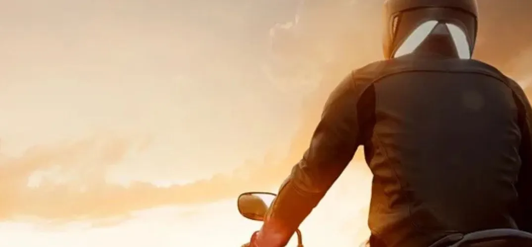 Person wearing a helmet and jacket riding a motorcycle at sunset with an orange sky background.