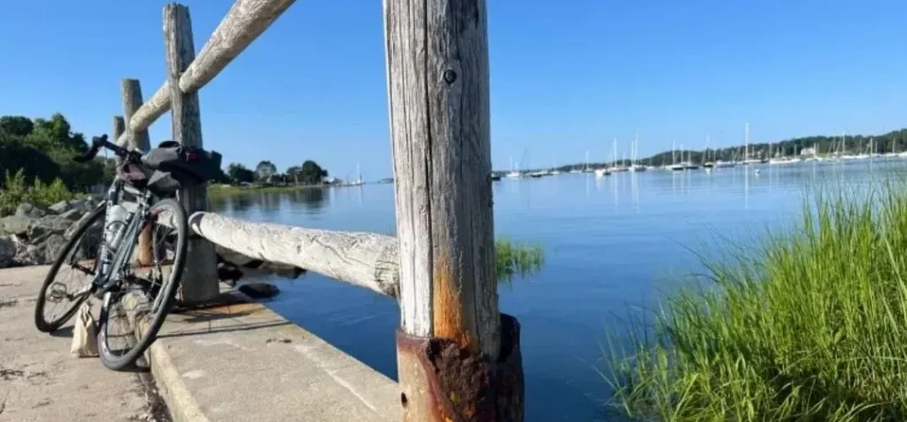 Bicycle leaning against a weathered wooden railing by a calm waterfront with sailboats in the distance.