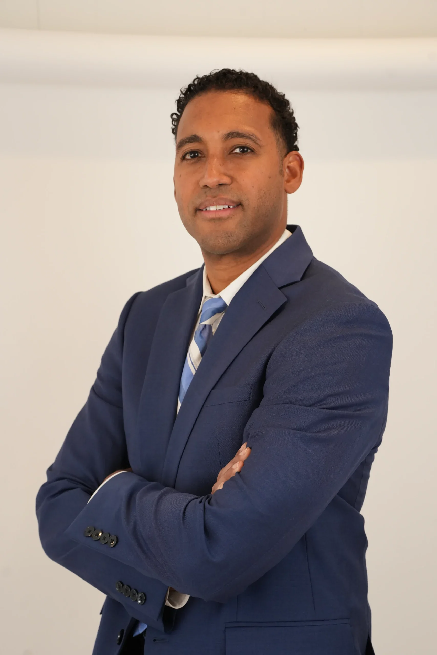 Professional man with folded arms wearing a navy blue suit and striped tie against a plain background.