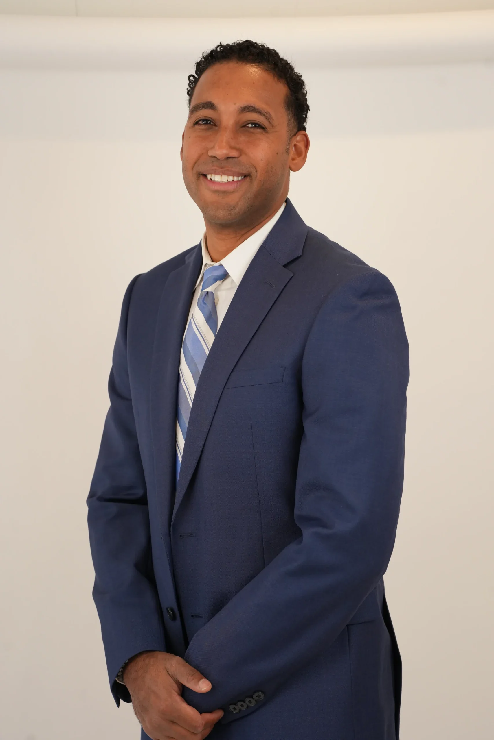Smiling man in a navy blue suit with a striped tie against a plain background.