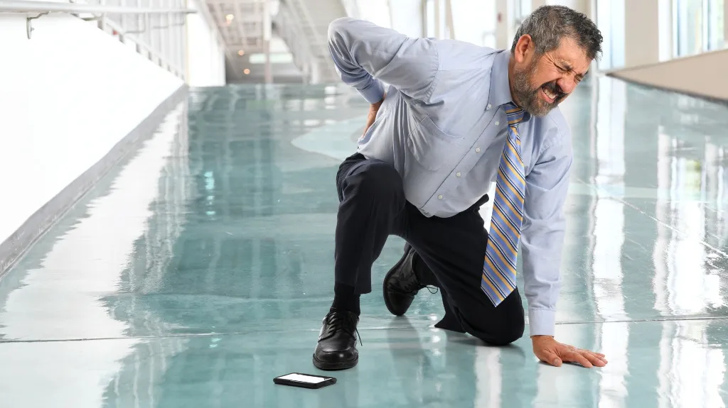 Middle-aged man in business attire kneeling on shiny floor, clutching lower back in pain, smartphone nearby.