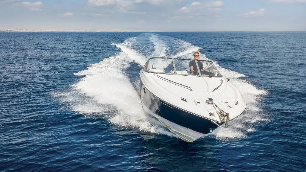 Person steering a white and navy speedboat on calm blue ocean waters under a partly cloudy sky.