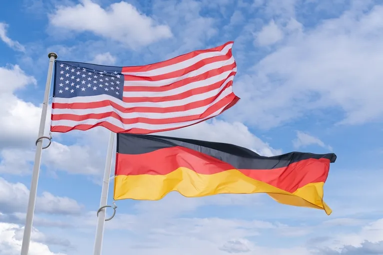 Two flags, United States and Germany, waving on flagpoles against a partly cloudy blue sky.
