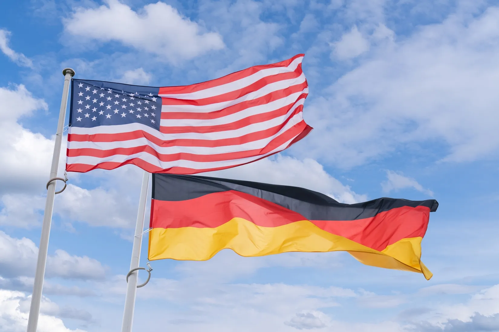 Flags of the United States and Germany waving on poles against a partly cloudy blue sky.