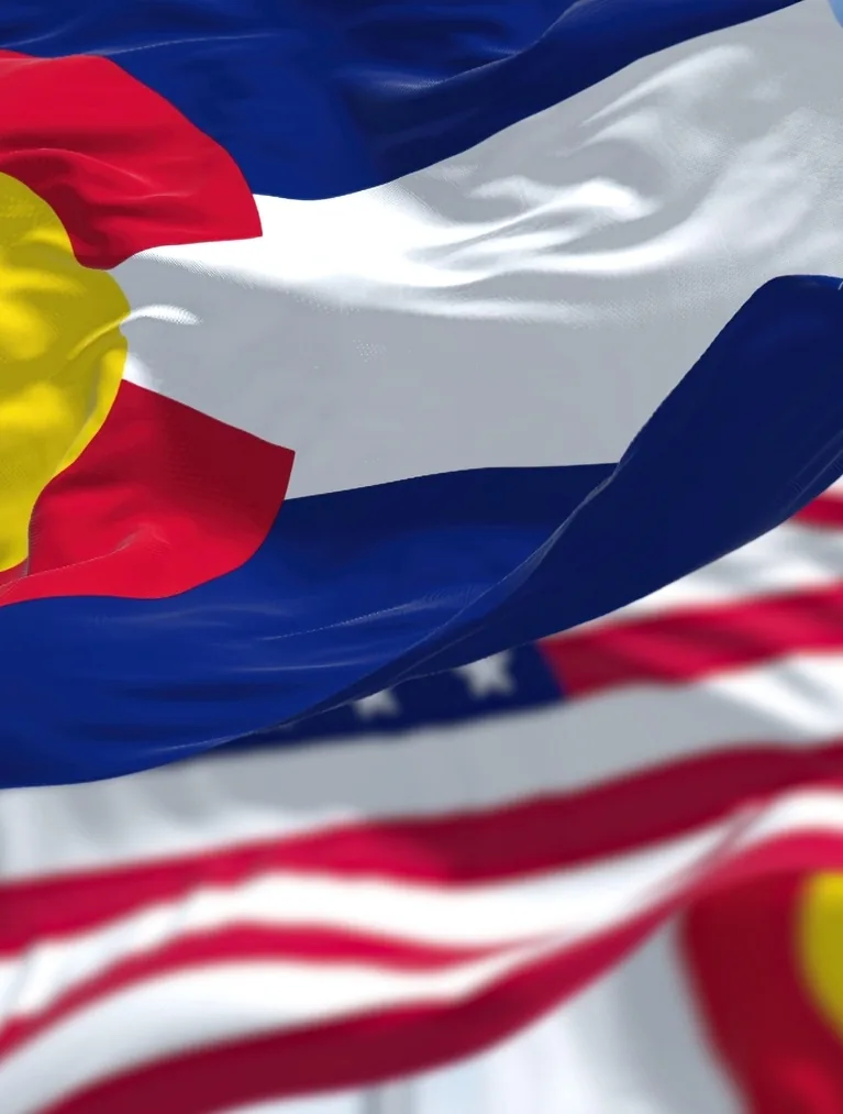 The Colorado state flag waving in the foreground with the United States flag behind it against a blue sky.
