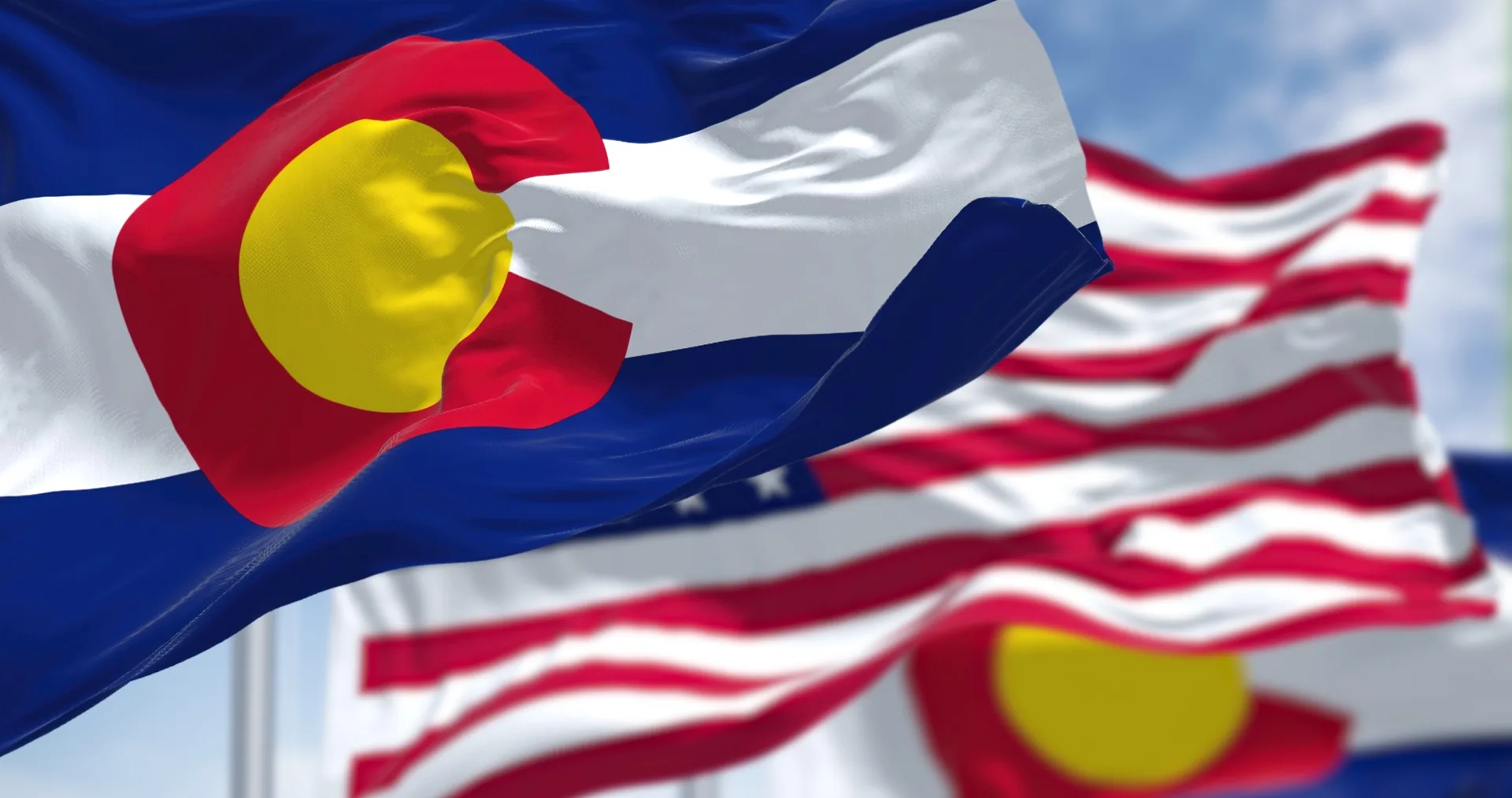The Colorado state flag waving in the foreground with the United States flag behind it against a blue sky.