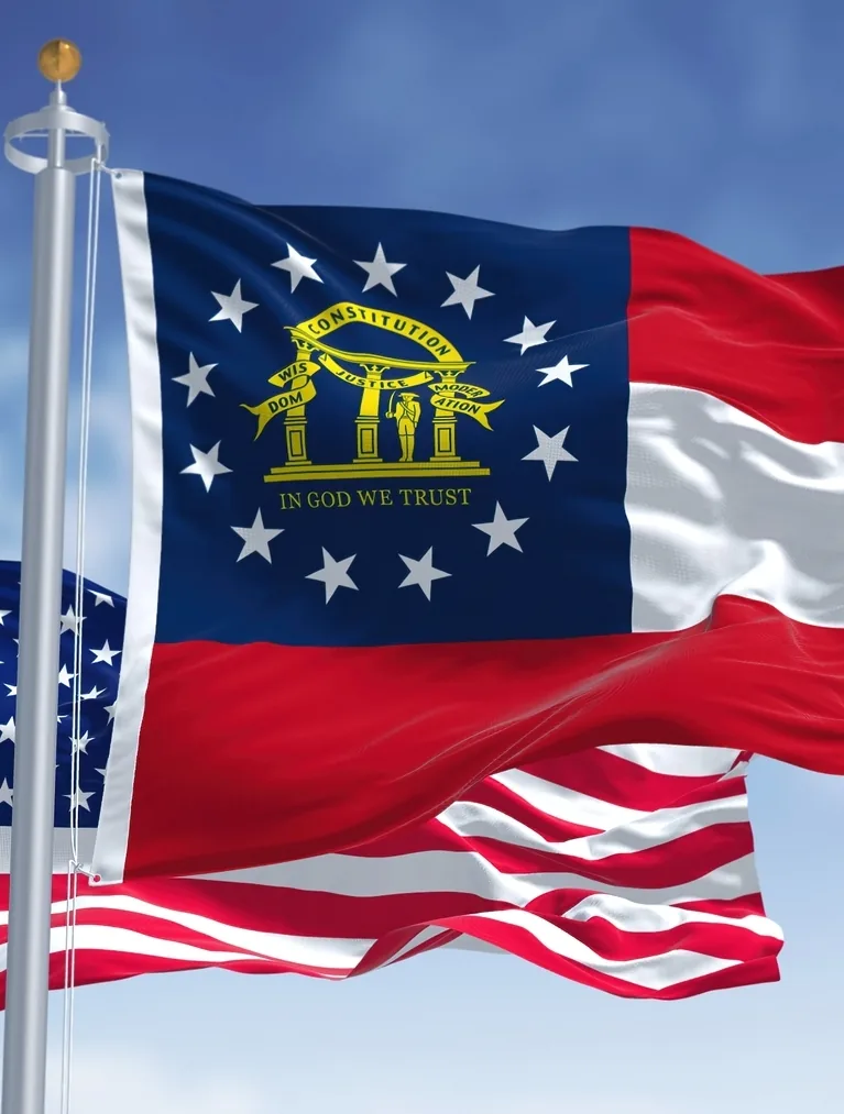 The Georgia state flag and the United States flag waving on flagpoles against a blue sky.