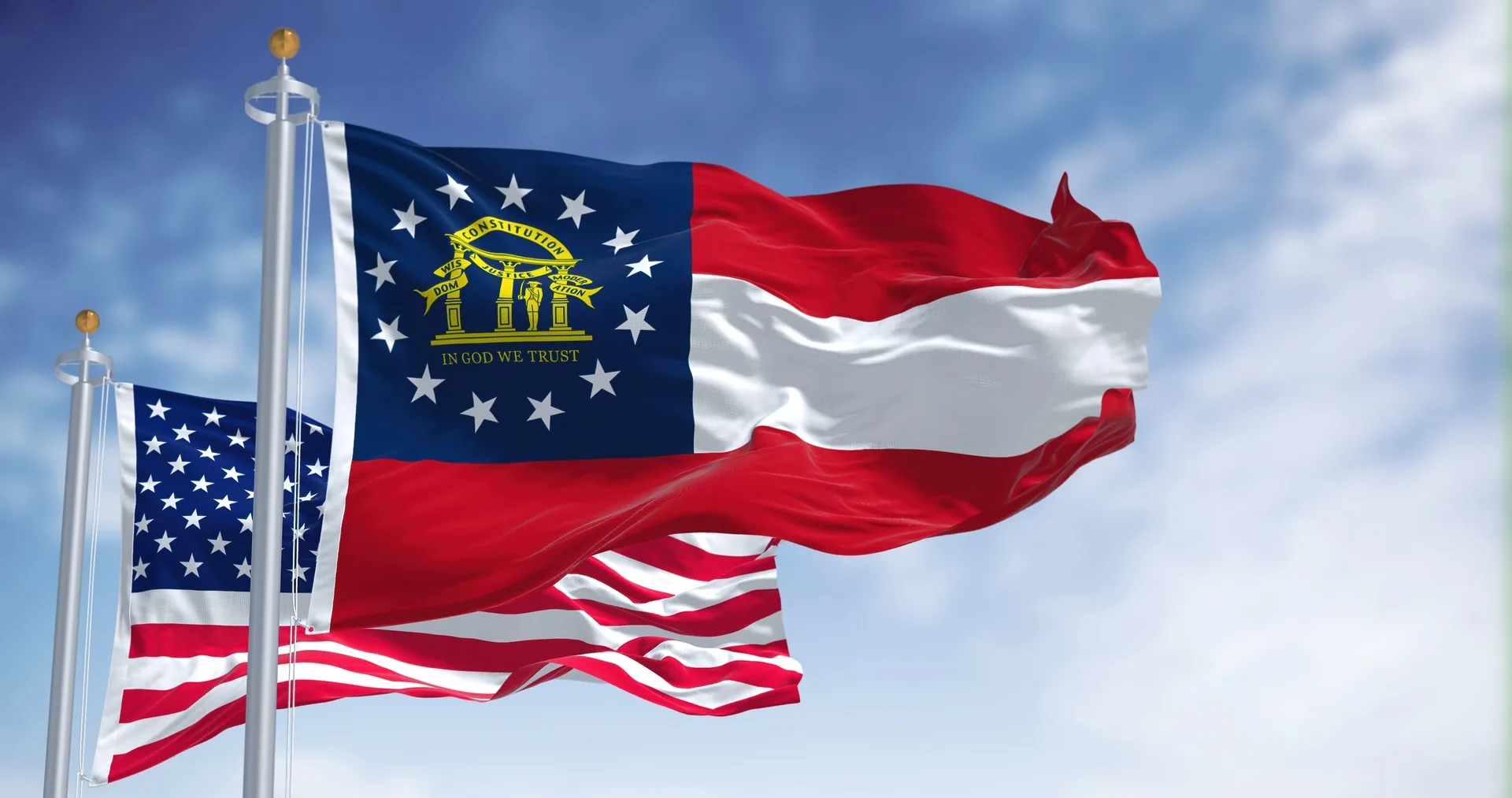 The Georgia state flag and the United States flag waving on flagpoles against a blue sky.
