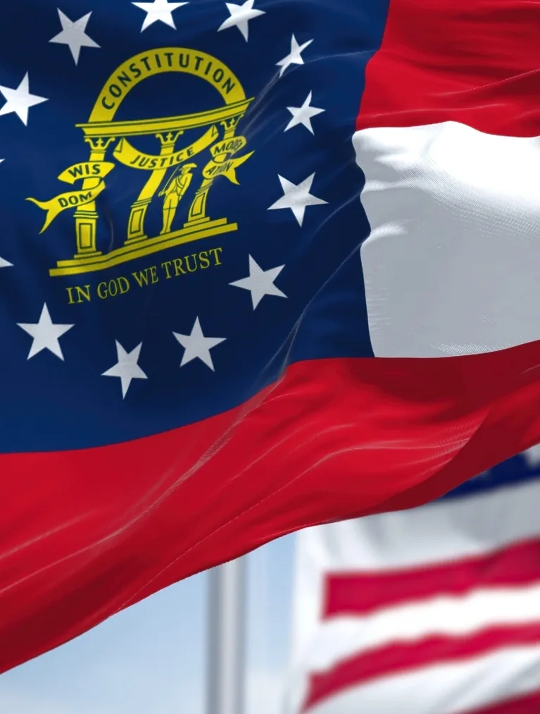 The Georgia state flag waving in the foreground with the United States flag behind it against a blue sky.