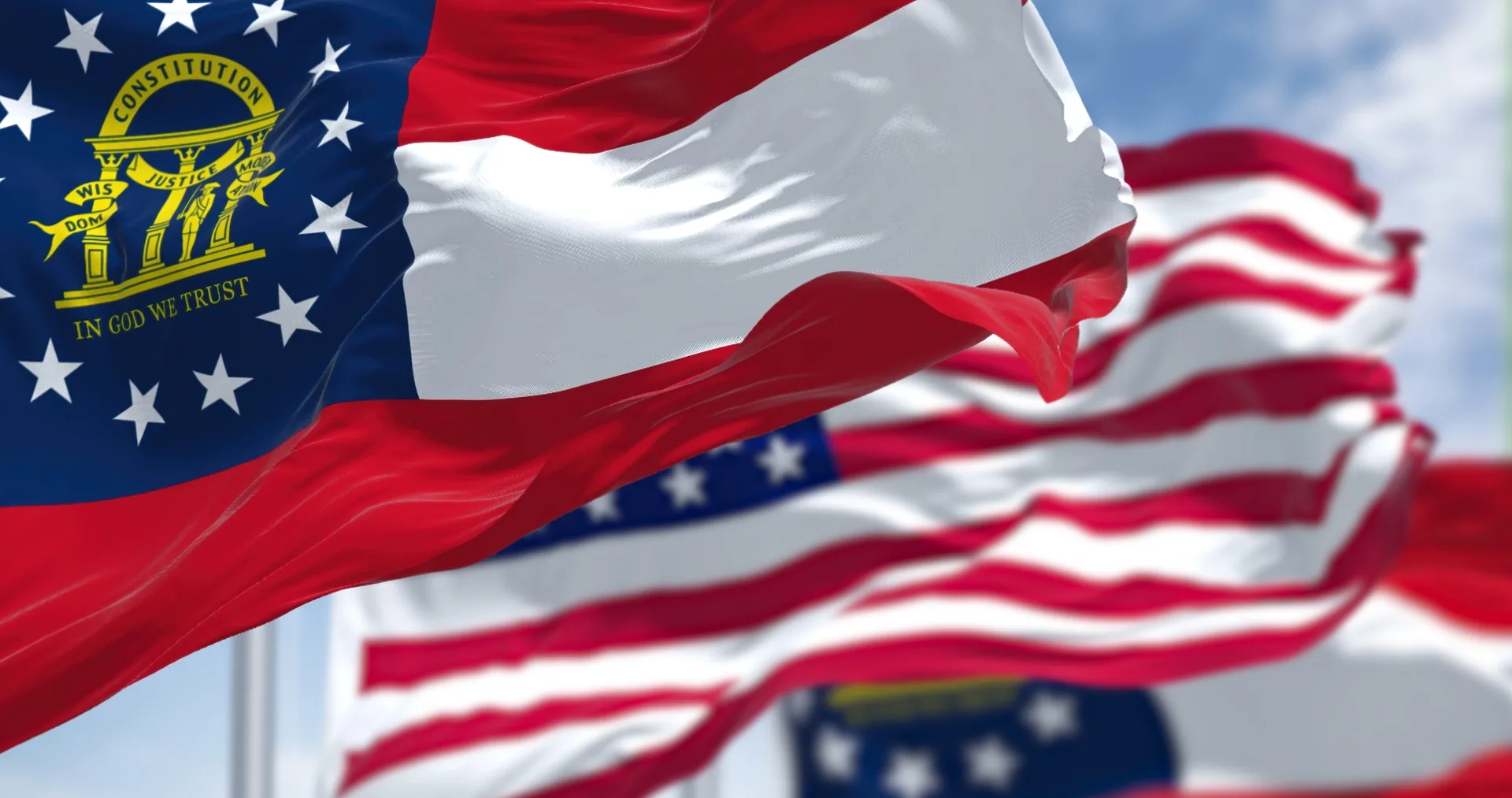 The Georgia state flag waving in the foreground with the United States flag behind it against a blue sky.