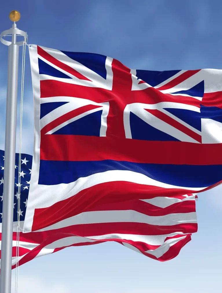 The Hawaii state flag and the United States flag waving on flagpoles against a blue sky.