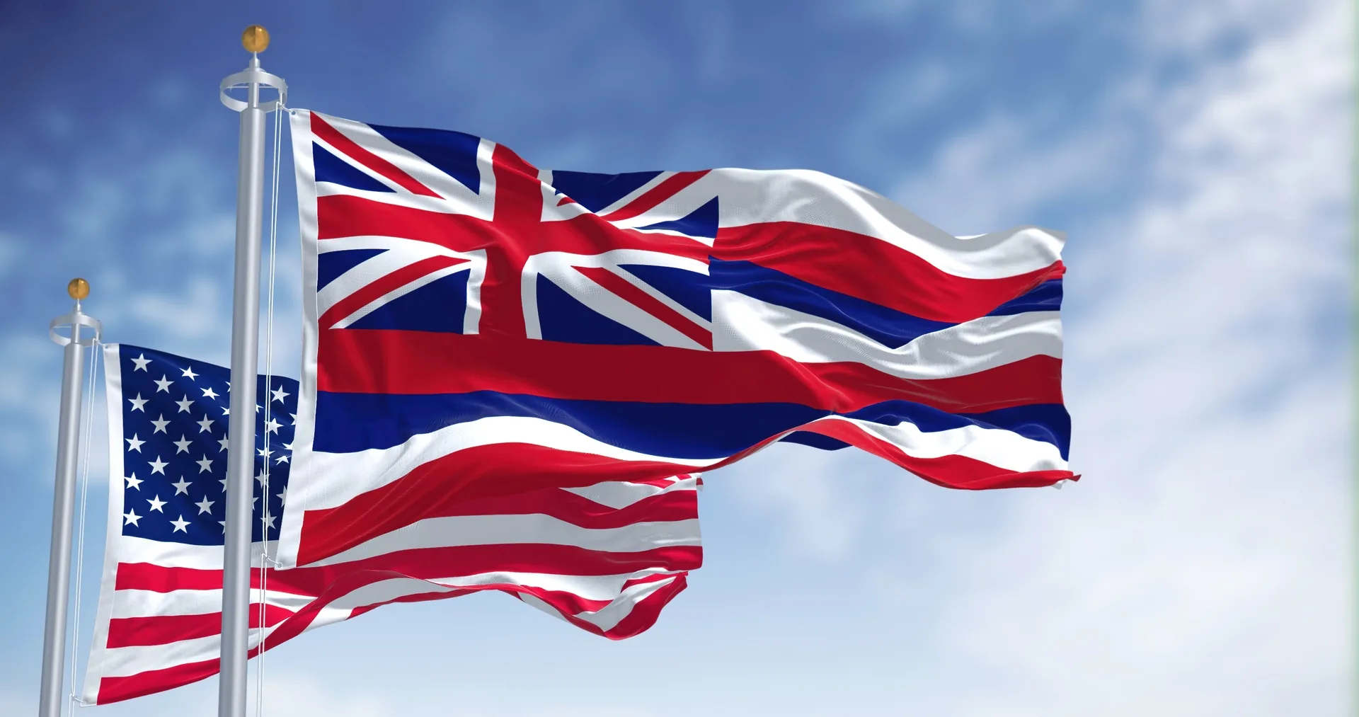 The Hawaii state flag and the United States flag waving on flagpoles against a blue sky.