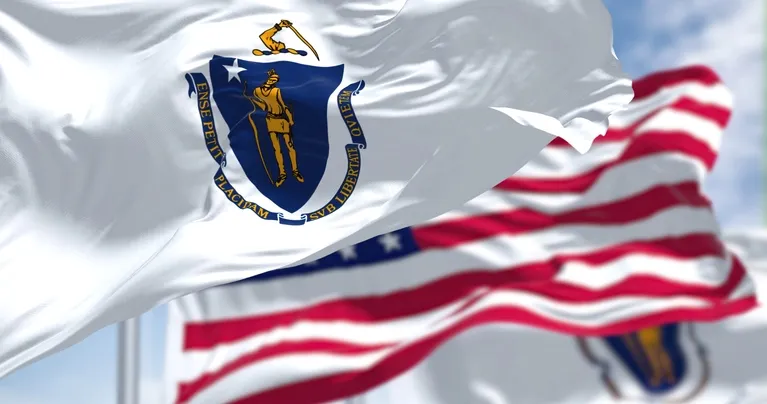 White Massachusetts state flag with blue shield and Native American figure, blurred American flag in background.