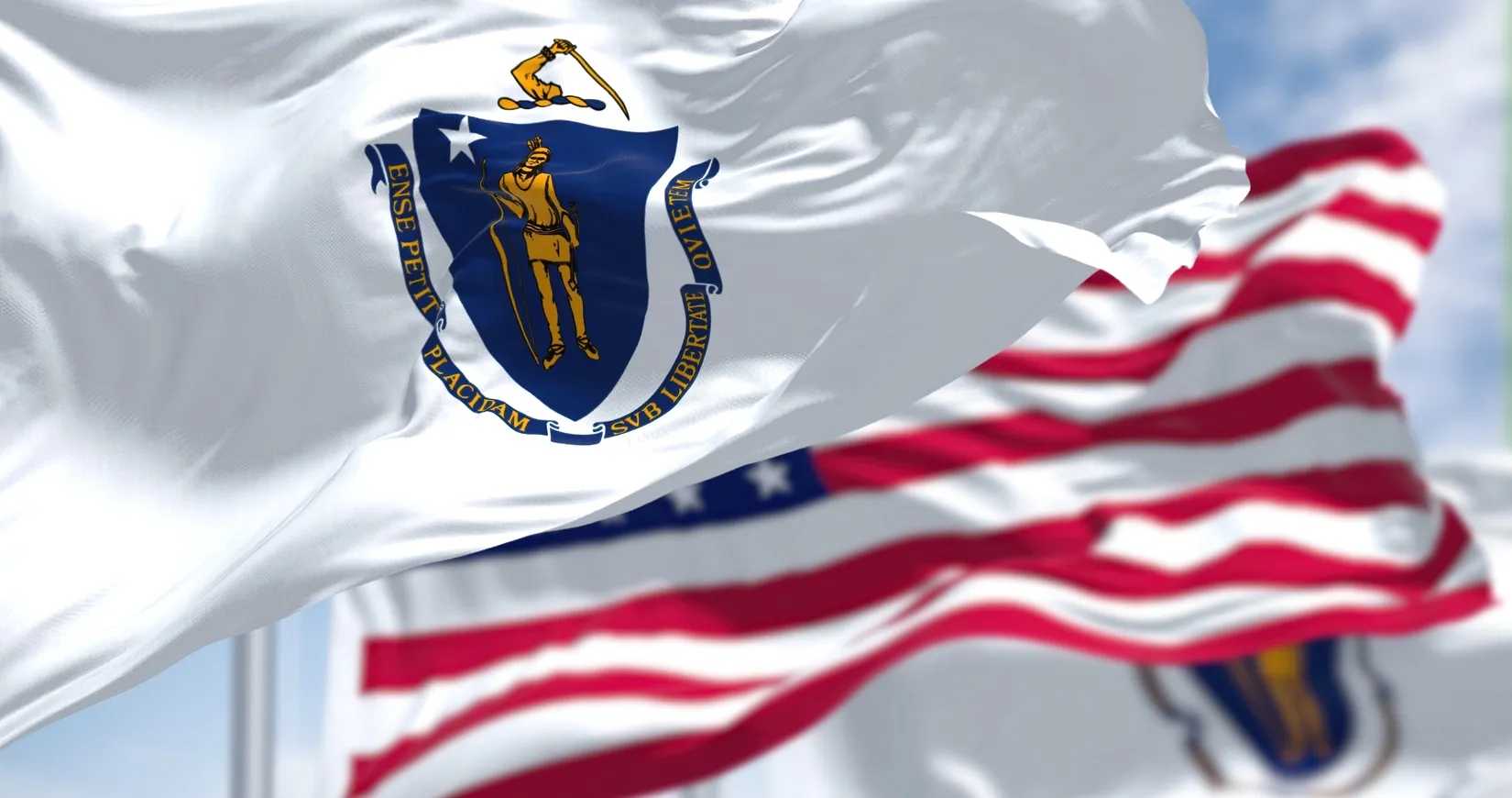 Waving Massachusetts state flag with blue shield and gold figure, American flag blurred in background.