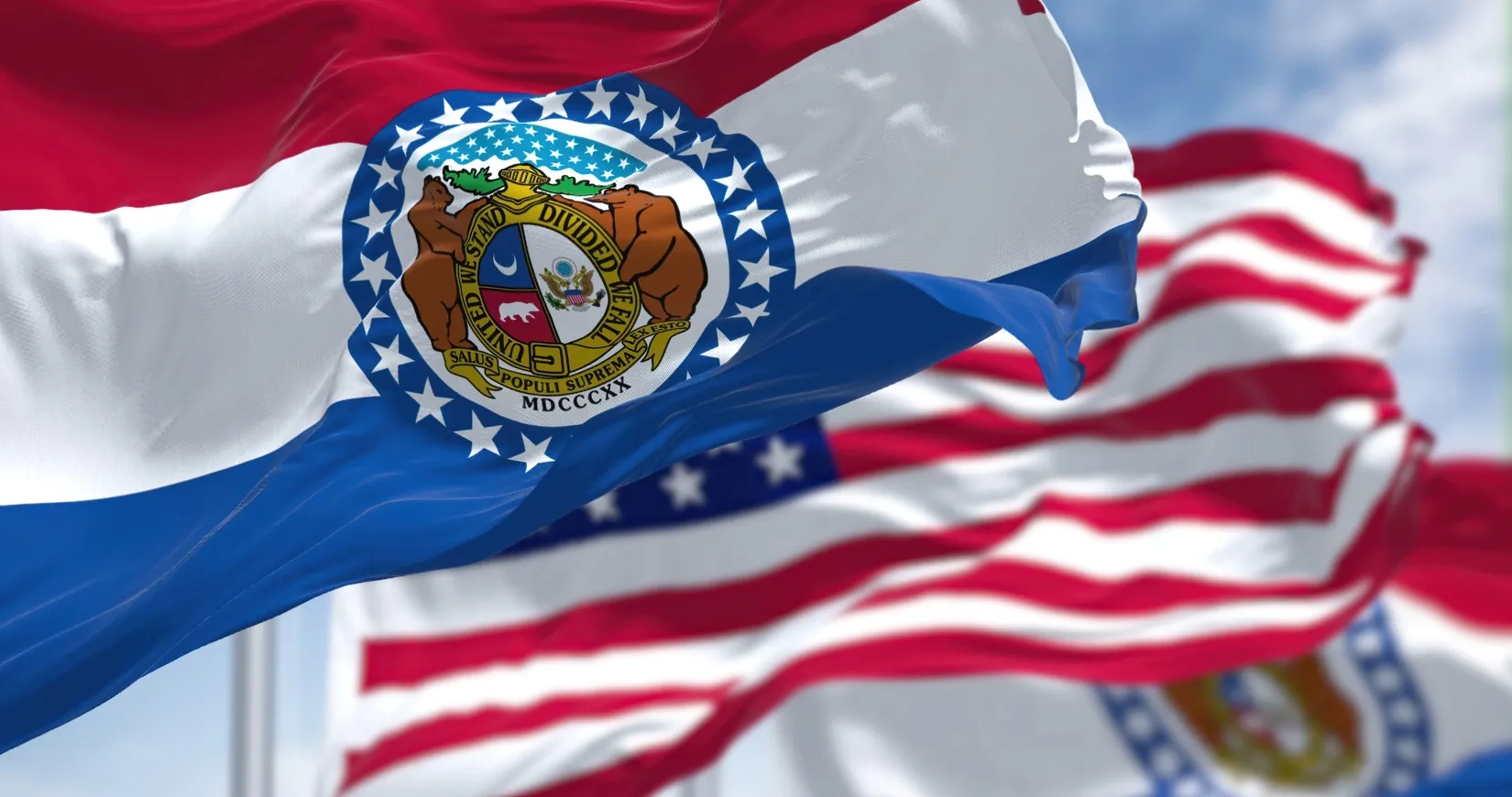 Waving Missouri state flag with detailed seal in foreground, American flags blurred in background.