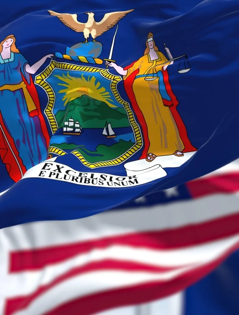 The New York state flag waving in the foreground with the United States flag behind it against a blue sky.