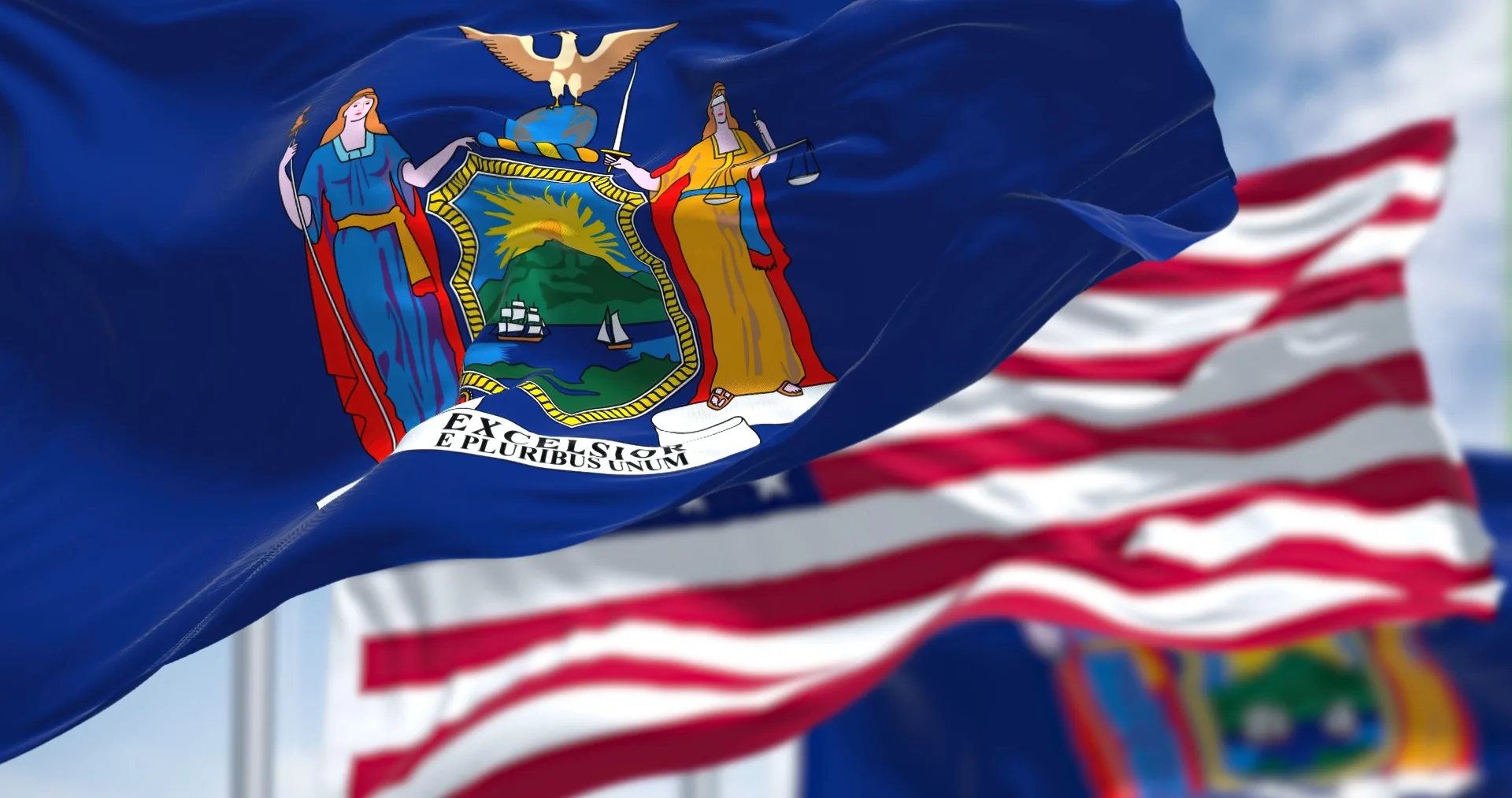The New York state flag waving in the foreground with the United States flag behind it against a blue sky.