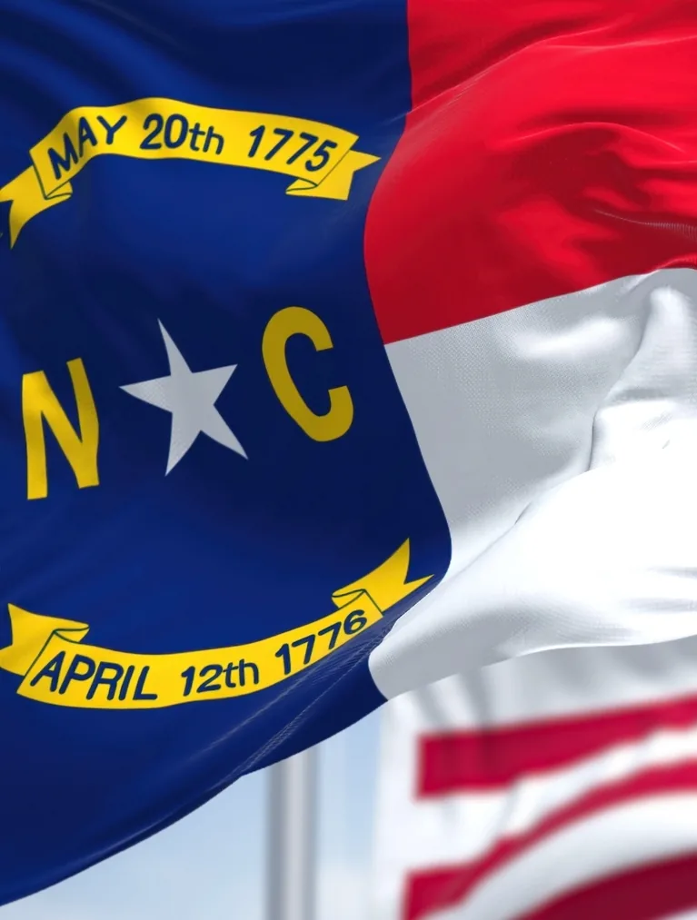 The North Carolina state flag waving in the foreground with the United States flag behind it against a blue sky.