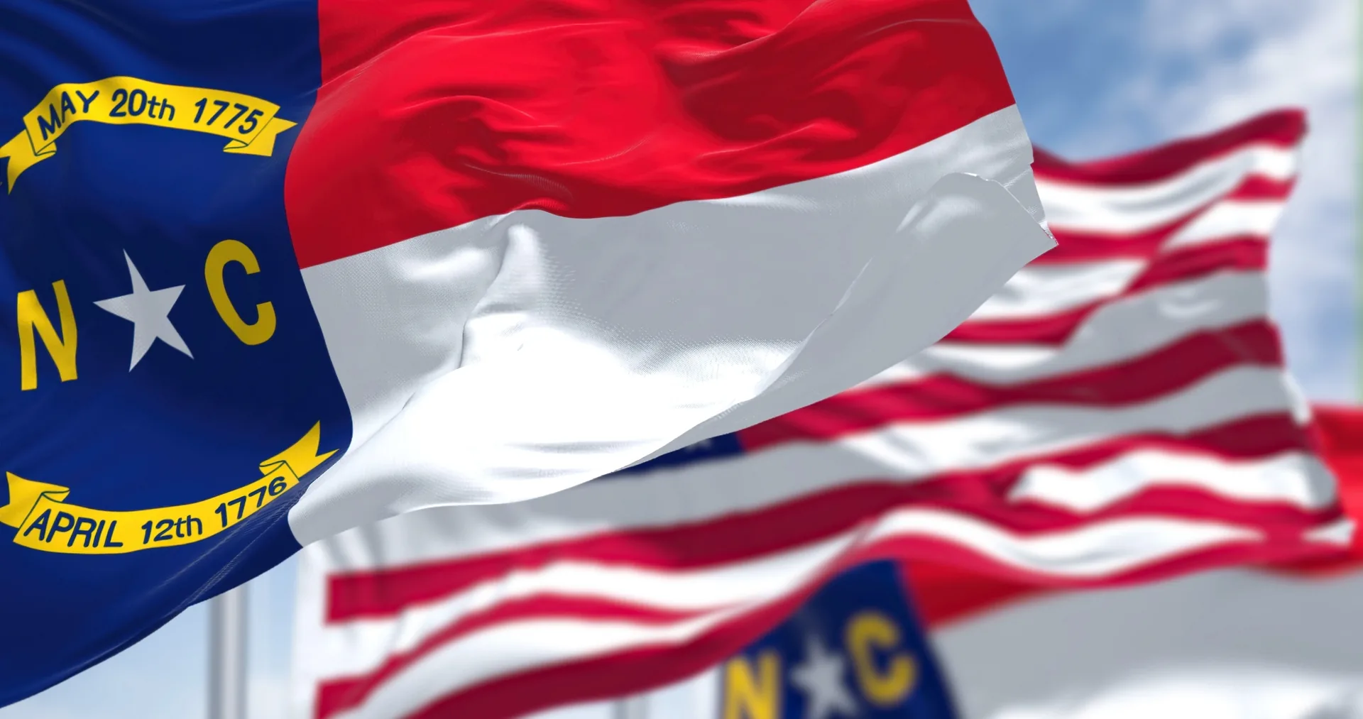The North Carolina state flag waving in the foreground with the United States flag behind it against a blue sky.