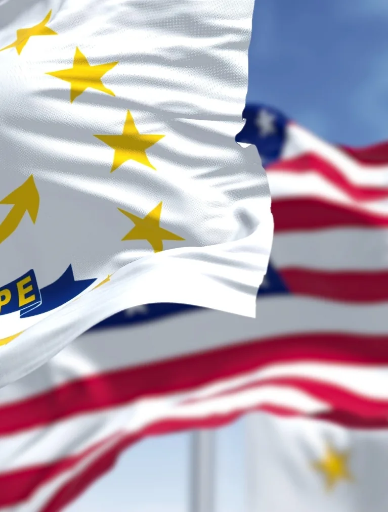 The Rhode Island state flag waving in the foreground with the United States flag behind it against a blue sky.