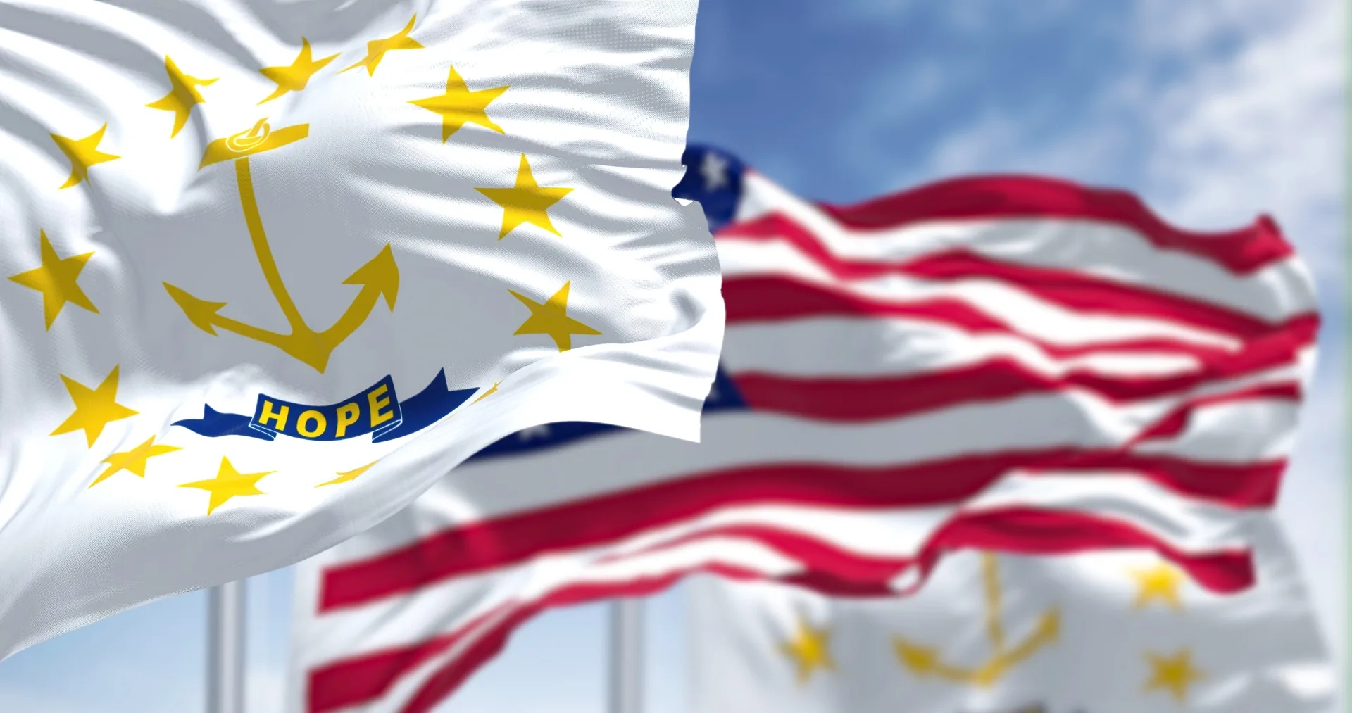 The Rhode Island state flag waving in the foreground with the United States flag behind it against a blue sky.
