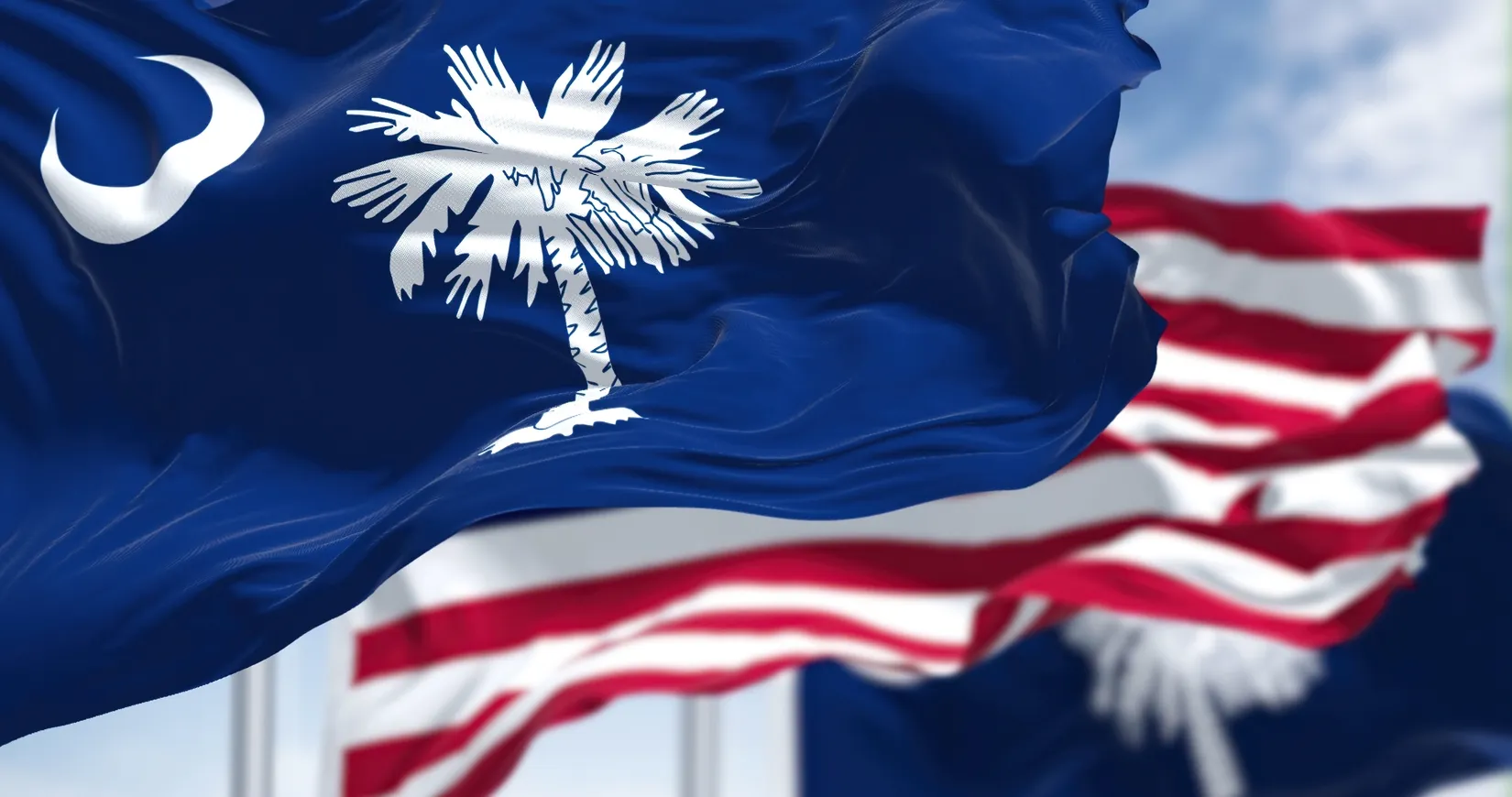 Waving South Carolina state flags with white palmetto and crescent on blue, American flags blurred in background.