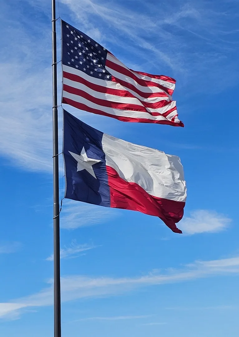 The United States flag and the Texas state flag flying on a single flagpole against a blue sky.
