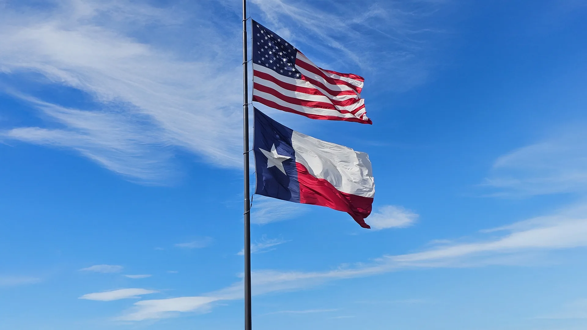 The United States flag and the Texas state flag flying on a single flagpole against a blue sky.