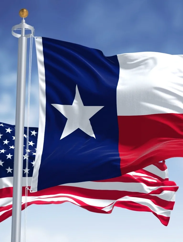 The Texas state flag and the United States flag waving on flagpoles against a blue sky.