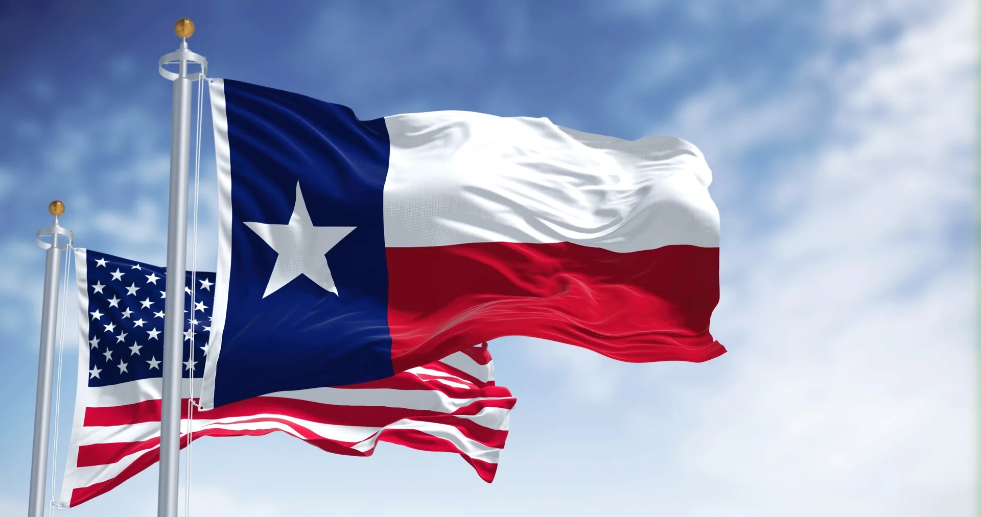 The Texas state flag and the United States flag waving on flagpoles against a blue sky.