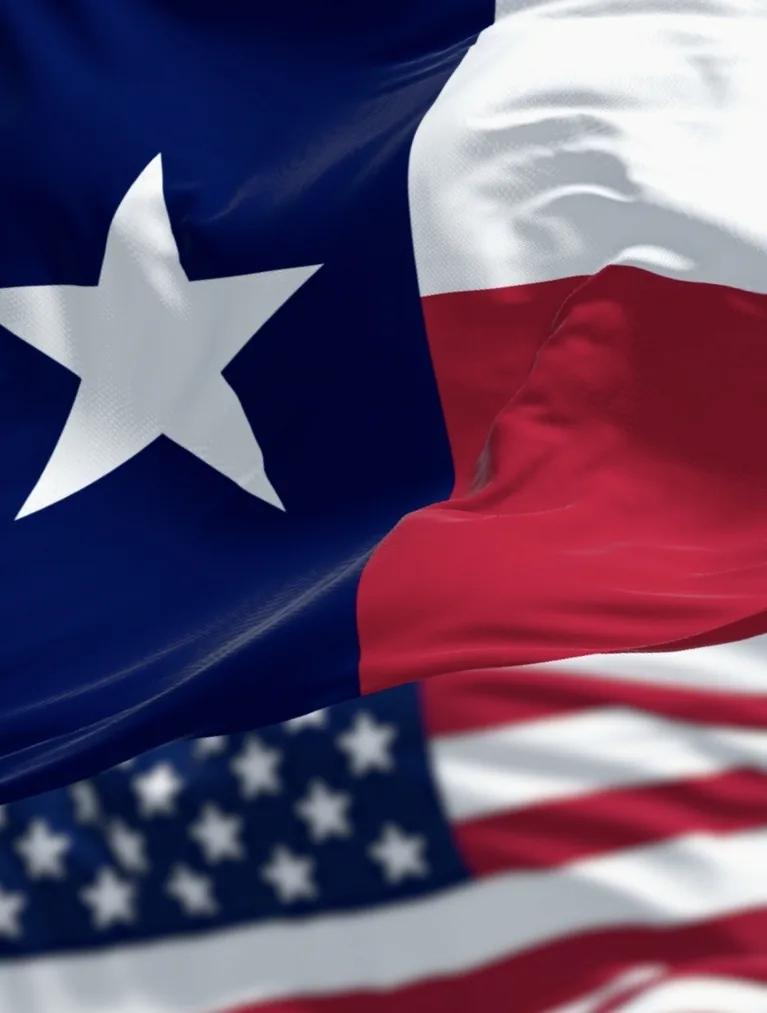 The Texas state flag waving in the foreground with the United States flag behind it against a blue sky.