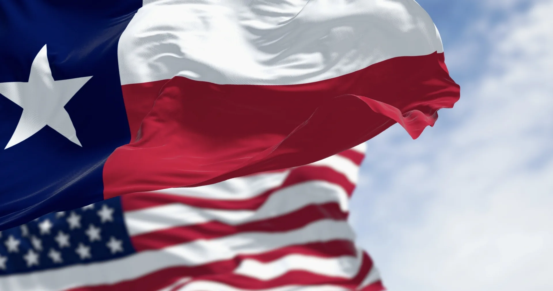 The Texas state flag waving in the foreground with the United States flag behind it against a blue sky.