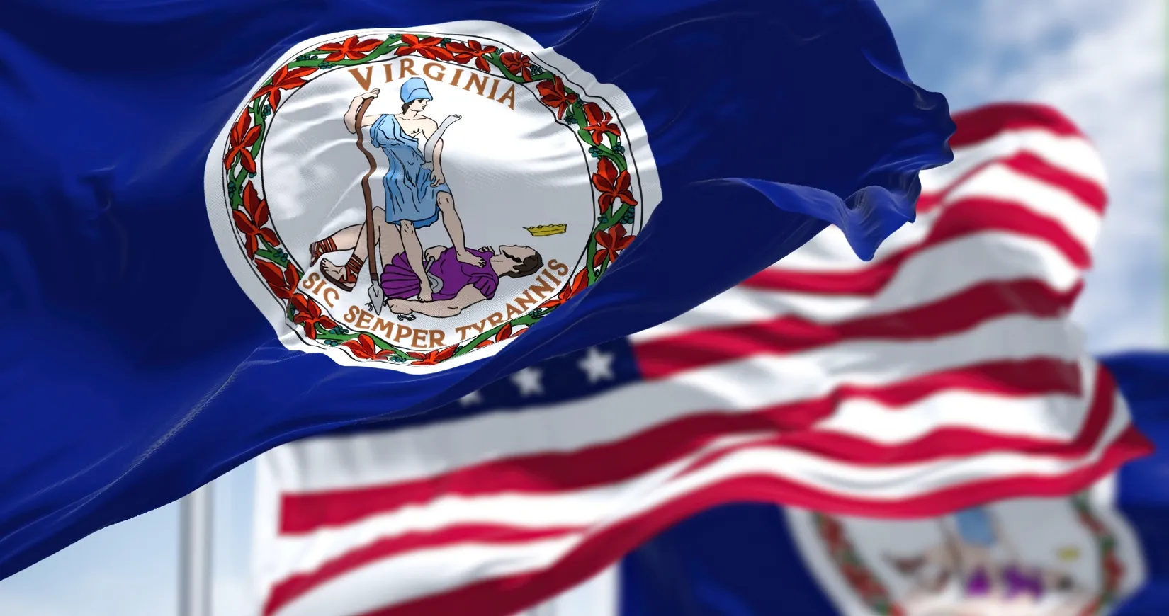 Waving Virginia state flag with seal and motto in the foreground, American flag in background.
