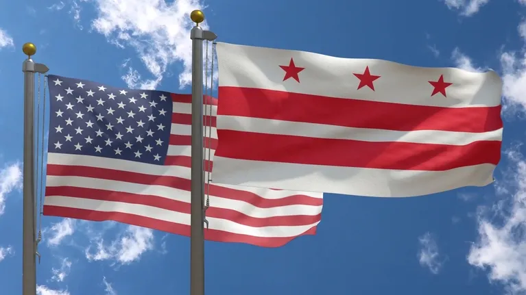 Two flags flying on flagpoles against a partly cloudy blue sky, including the United States flag and the Washington, D.C. flag.