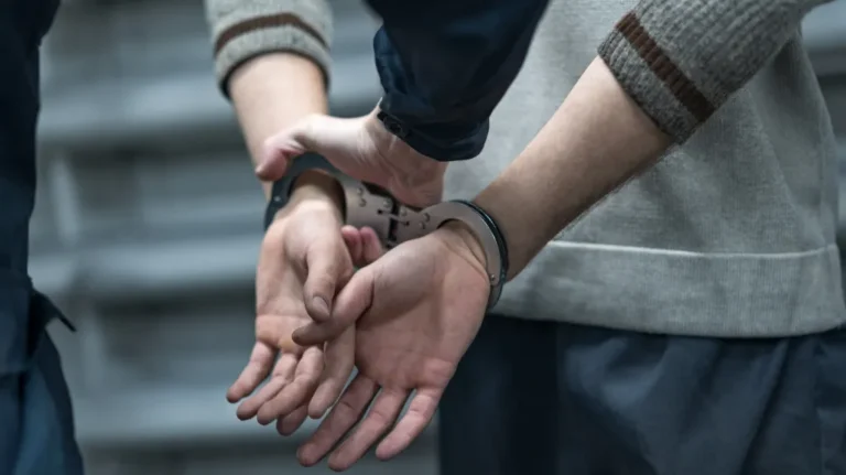 Close-up of a person in a gray sweater being handcuffed behind their back by another individual.