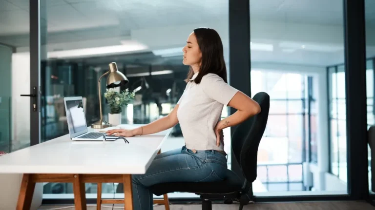Young woman in white shirt and jeans sitting at desk, holding lower back in pain.