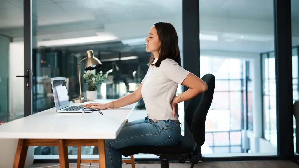 Young woman in white shirt and jeans sitting at desk, holding lower back in pain.