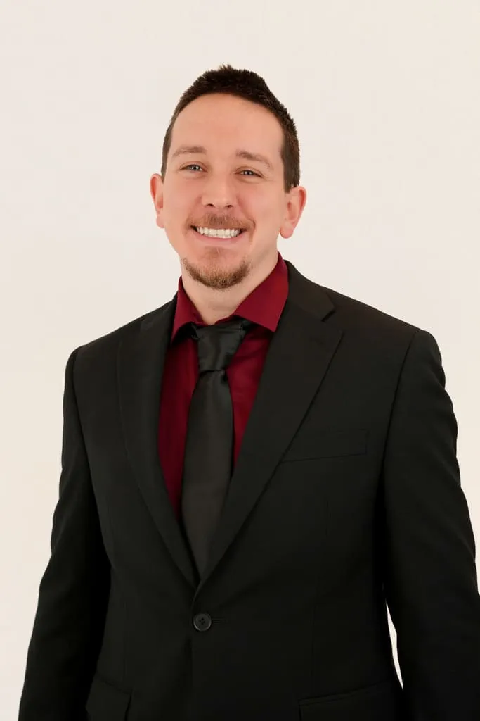 Portrait of a smiling man wearing a black suit, burgundy shirt, and black tie against a plain background.
